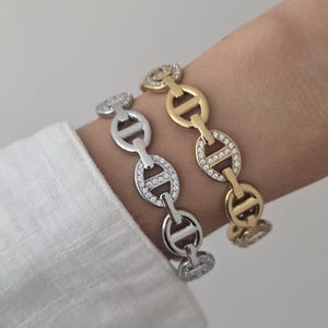 Peut inclure: Deux bracelets à maillons, l'un argenté et l'autre doré, composés de maillons ovales avec une barre horizontale. Certains maillons de chaque bracelet sont incrustés de petits cristaux ronds et transparents qui captent la lumière.