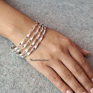 May include: A set of three silver-toned bangles featuring a continuous twisted, spiral design. The bracelets are worn together on the wrist, showcasing a polished metallic finish with uniform curves that create a rhythmic, repeating pattern.