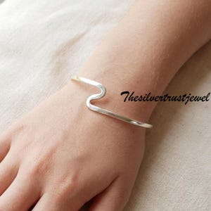 Pode incluir: Uma pulseira de prata polida com uma curva ondulada distinta no centro da banda. O metal tem um acabamento liso e reflexivo. O texto Thesilvertrustjewel é visível em letras pretas à direita da pulseira.