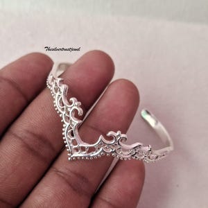 Puede incluir: Una pulsera de brazalete de plata con un intrincado diseño en forma de V puntiaguda con delicados recortes de filigrana y un borde de cuentas. El metal tiene un acabado pulido y reflectante. El texto Thesilvertrustjewel es visible en la esquina superior izquierda.