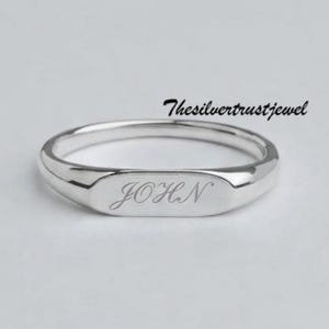 Può includere: Un anello color argento lucido con una faccia piatta rettangolare incisa con il nome JOHN in corsivo. Il testo Thesilvertrustjewel è posizionato sopra l'anello. La fascia è liscia e presenta un profilo arrotondato e uniforme.