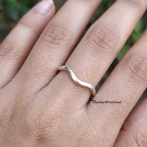 Puede incluir: Un anillo de plata pulida que presenta una banda lisa y continua en forma de onda. El metal tiene un acabado metálico reflectante que se curva suavemente sobre el dedo. El texto Thesilvertrustjewel está impreso en pequeñas letras negras.