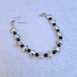 Peut inclure: Un bracelet argenté composé d'une alternance de perles noires et de petites breloques en forme de tortue. Le bracelet comprend une chaînette d'extension en métal avec un fermoir mousqueton. Le texte Thesilvertrustjewel est imprimé près du fermoir.
