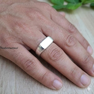 Puede incluir: Un anillo de sello de plata pulida con una superficie superior plana y rectangular. La banda es sólida y metálica, con un acabado limpio y reflectante que rodea el dedo. El diseño es sencillo, presentando una cara lisa sin grabados.