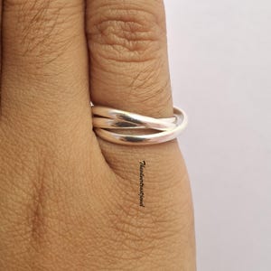 Può includere: Un anello a tripla fascia in argento lucido composto da tre anelli metallici lisci e intrecciati che si avvolgono attorno al dito. L'anello ha una finitura metallica riflettente e include il piccolo testo nero Thesilvertrustjewel stampato verticalmente.