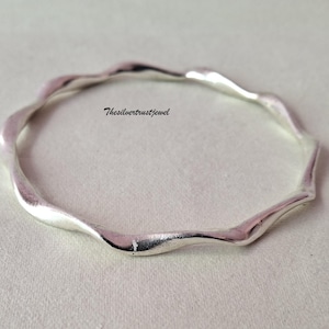 May include: A polished silver bangle bracelet featuring a continuous wavy, undulating design. The metal surface is smooth and reflective, catching the light along its curved edges. The text Thesilvertrustjewel is printed in black in the center.