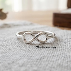 May include: A polished silver ring featuring a central infinity symbol design. The band is smooth and circular, showcasing a continuous loop shape that represents the mathematical sign for infinity. The text Thesilvertrustjewel is visible.