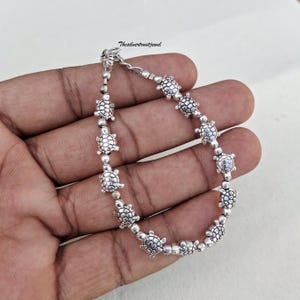 Könnte beinhalten: Ein silberfarbenes Armband mit einer Reihe kleiner, detaillierter Schildkrötenanhänger, die durch runde Perlen miteinander verbunden sind. Das Metall hat eine polierte Oberfläche mit dem Text Thesilvertrustjewel nahe dem Verschluss.