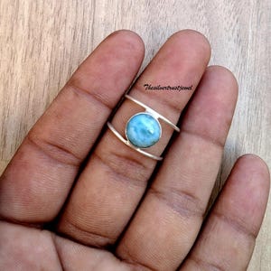 Puede incluir: Un anillo de plata con una piedra preciosa Larimar redonda de color azul claro engastada entre dos bandas de plata paralelas. El metal tiene un acabado pulido y el texto Thesilvertrustjewel es visible en letras negras pequeñas arriba.
