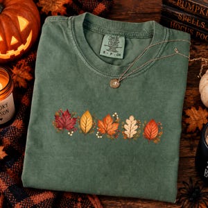 May include: A sage green short-sleeve t-shirt featuring a row of five colorful autumn leaves printed across the chest. The shirt has a crew neckline and a Comfort Colors brand label on the inside collar. A thin gold chain necklace rests on the fabric.