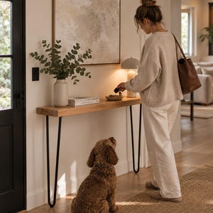 Puede incluir: Una mesa consola de madera larga y estrecha con patas de horquilla de metal negro. La mesa sostiene un jarrón de cerámica beige con ramas de eucalipto verdes, un libro blanco, un cuenco pequeño con llaves y una lámpara beige redonda.