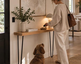 Peut inclure: Une console en bois longue et étroite avec des pieds en épingle en métal noir. La table supporte un vase en céramique beige avec des branches d'eucalyptus vertes, un livre blanc, un petit bol avec des clés et une lampe beige ronde.