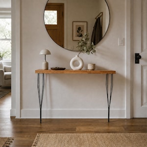 Puede incluir: Una mesa consola de madera con acabado natural y cuatro patas finas de metal negro. Sobre la mesa hay una pequeña lámpara beige, un cuenco oscuro poco profundo, un jarrón de cerámica blanco en forma de donut con follaje verde y un pequeño recipiente cilíndrico beige.