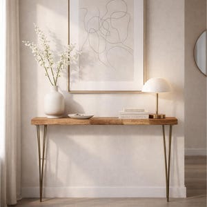 Pode incluir: Uma mesa consola de madeira com pernas de metal finas, com um vaso branco contendo flores brancas, uma pequena tigela rasa, uma pilha de livros e um candeeiro em forma de cogumelo. Acima da mesa, uma impressão de arte linear emoldurada.
