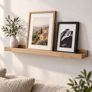 May include: A light oak wood floating ledge shelf holding a white ceramic vase with green leaves, a large framed landscape print of a hillside village, and a smaller black framed portrait photograph of two people in black and white.