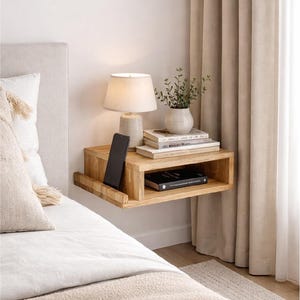 Puede incluir: Una mesita de noche flotante de madera clara montada en la pared. El estante rectangular cuenta con un compartimento de almacenamiento abierto, una ranura lateral para un teléfono inteligente y una superficie superior con una lámpara y libros.