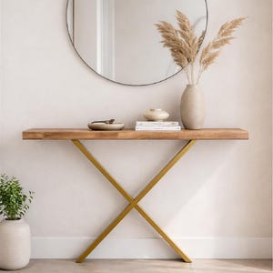 Op de afbeelding: Een rechthoekige houten sidetable met een natuurlijke houtnerfafwerking en een goudkleurig metalen X-vormig onderstel. Op het tafeloppervlak staan een kleine keramische kom, twee gestapelde boeken en een beige vaas met gedroogd pampasgras.