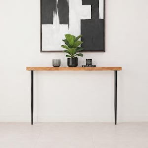 May include: A rectangular wooden console table with a natural grain finish and four slender black metal legs. On the table sits a green potted plant in a black container, flanked by a ribbed gray glass vessel and a small black textured candle holder.