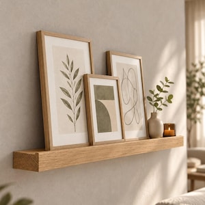 Può includere: Una mensola sospesa in rovere chiaro che sostiene tre stampe incorniciate con disegni botanici e astratti, un piccolo vaso in ceramica beige contenente un rametto verde e un portacandele in vetro scuro con una candela accesa su una parete neutra.