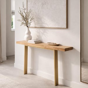May include: A rectangular console table featuring a thick, natural light-toned wooden top supported by two flat, vertical gold-colored metal legs. The table surface holds a white ceramic vase with dried stems, a stack of books, and a small oval dish.