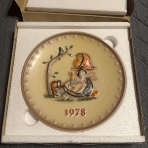 May include: A round ceramic collector plate featuring a raised relief illustration of a child sitting under a tree with a bird. The plate has a cream-colored background, a brown border with embossed stars, and the red printed year 1978 at the bottom.