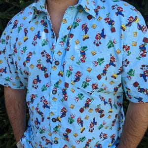 May include: A light blue short-sleeved button-down shirt with a repeating pattern of Mario characters, mushrooms, and question blocks.