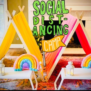 May include: Two colorful teepees with rainbow pillows and white trays with lanterns. The teepees are made of wood and fabric. The text "SOCIAL DISTANCING CAMP" is written in green letters on a yellow banner.