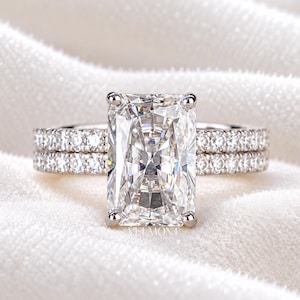 May include: A silver-toned ring set featuring a large, rectangular radiant-cut center stone held by four prongs. The band and the accompanying stackable band are both encrusted with small, round, clear pavé stones along the entire visible surface.