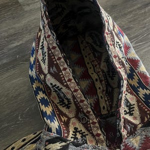 Op de afbeelding: Een hoodie met rits voorzien van een geweven tapijtachtig patroon in de kleuren donkerrood, marineblauw, zwart en crème. Het kledingstuk heeft een capuchon met een bijpassend ingewikkeld geometrisch ontwerp en een metalen ritssluiting.