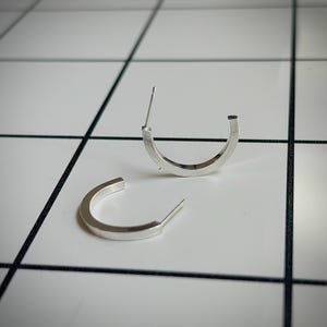 以下が含まれることがあります： 平らな半円形デザインのシルバーカラーのフープピアス一対。各ピアスには片端から伸びる細いポストが付いており、ピアスホールに通す仕様です。金属は滑らかで光沢のある仕上げで、全体を通して均一な厚みがあります。