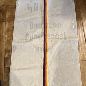 May include: A vintage, off-white canvas mail sack with a black, red, and yellow stripe down the center. The sack has the text "Deutsche Bundespost" printed on it.