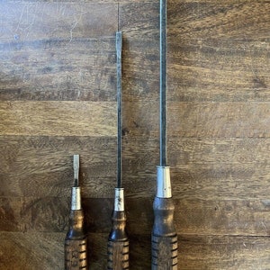 May include: Three vintage screwdrivers with wooden handles and metal shafts. The handles are brown with yellow accents. The screwdrivers are arranged in order of size, with the largest screwdriver on the right and the smallest screwdriver on the left.