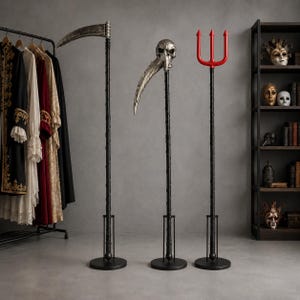 May include: Three tall props standing on circular black bases: a scythe with a curved silver blade, a staff topped with a silver skull and curved blade, and a trident with a bright red three-pronged head, all featuring black textured shafts.