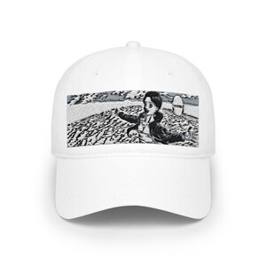 May include: A white baseball cap featuring a rectangular black and white graphic print on the front panel. The illustration depicts a person with dark hair and an open jacket standing with arms outstretched against a textured, landscape background.
