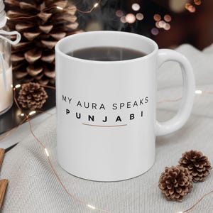 以下が含まれることがあります： 前面に黒い文字で「MY AURA SPEAKS PUNJABI」とプリントされた白いセラミック製のマグカップ。マグカップには標準的な持ち手があり、中には濃い色の液体が入っています。デザインにはPunjabiという単語の下に細い水平線が含まれています。