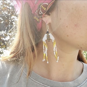 May include: A pair of dangle earrings featuring a beaded bee design with black, white, and gold accents. The earrings are made with small beads and have a long, flowing design.