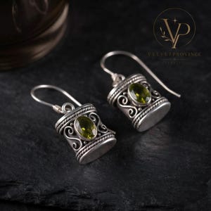 May include: A pair of sterling silver drop earrings featuring a cylindrical design with intricate scrollwork and twisted wire detailing. Each earring is set with a single oval-cut, faceted olive-green gemstone and includes a curved silver ear wire.