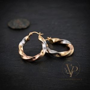 May include: A pair of hoop earrings featuring a twisted design with alternating sections of yellow gold, rose gold, and silver-toned metal. The earrings have a hinged post closure mechanism and a polished, reflective finish on the metallic surfaces.