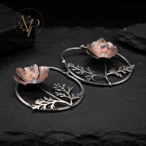 Puede incluir: Un par de pendientes de aro circulares que presentan una flor de metal en tonos cobre y un tallo de metal plateado oscuro oxidado con follaje detallado. El diseño floral está centrado dentro del aro fino, creando una estética botánica.