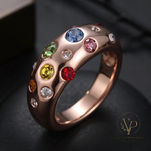 May include: A rose gold-toned ring featuring a wide, smooth band with a polished finish. The surface is embedded with multiple round, multi-coloured gemstones including blue, green, yellow, red, orange, pink, and clear stones set in a scattered pattern.