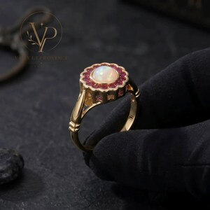 May include: A gold ring featuring a central round cabochon opal surrounded by a halo of small, round, deep pink rubies. The setting has a scalloped edge design, and the band is smooth and polished, showcasing a classic, refined metal construction.
