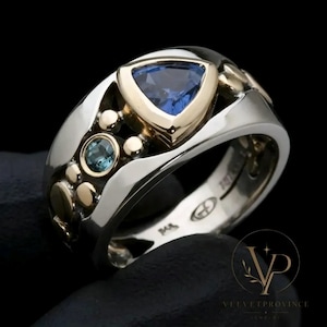 Op de afbeelding: Een tweekleurige ring met een grote, in trillion geslepen blauwe edelsteen gezet in geelgoud, geaccentueerd door een kleinere ronde blauwe edelsteen en diverse gepolijste geelgouden bolletjes in een brede, gladde zilveren ringband.