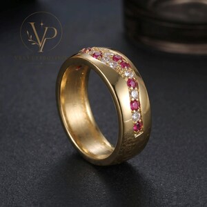 May include: A gold-toned band ring featuring a central channel set with alternating round red and clear stones. The metal surface has a smooth finish on the sides and a textured, hammered-like appearance on the lower portion of the band.