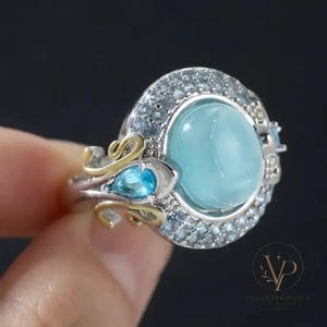 Könnte beinhalten: Ein silberner Ring mit einem großen, ovalen, hellblauen Cabochon-Stein in der Mitte, umgeben von einem Kranz aus kleinen, funkelnden blauen Steinen. Die Ringschiene zeigt goldfarbene Schnörkel und einen birnenförmigen blauen Stein an der Seite.