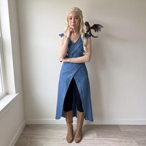 Könnte beinhalten: Ein blaues ärmelloses Kleid mit V-Ausschnitt und Wickelrock, getragen über einer schwarzen Leggings und hellbraunen Stiefeln. Das Outfit umfasst eine lange, geflochtene blonde Perücke und eine kleine braune Drachenfigur auf der linken Schulter.