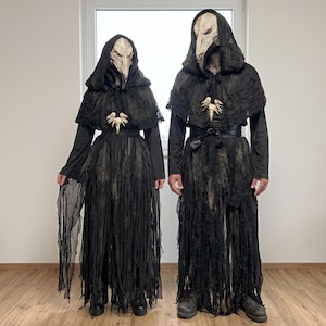 May include: Two figures wearing black hooded cloaks with distressed, shredded fabric layers and long, pointed beige bird-like masks. Each outfit includes a black base garment, a waist belt, and a necklace featuring a small, carved beige bird skull pendant.