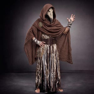 Può includere: Un mantello marrone con cappuccio indossato sopra un indumento a strati beige e marrone con una cintura a corsetto marrone scuro. L'outfit include una maschera da medico della peste, fasce per le braccia abbinate e una gonna con strisce di tessuto.