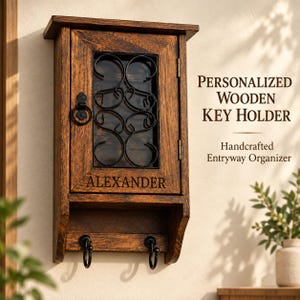 Könnte beinhalten: Ein dunkelbrauner, wandmontierter Schlüsselkasten aus Holz mit einer Flügeltür, die einen schwarzen Metalleinsatz mit Schnörkeln und einen Ringgriff aufweist. Der Name ALEXANDER ist in die Tür eingraviert. Darunter befinden sich zwei Haken.