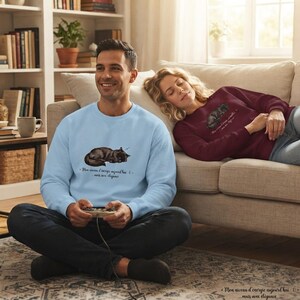 May include: A light blue crewneck sweatshirt featuring a graphic of a sleeping black cat and the French text "Mon niveau d'énergie aujourd'hui : 0... mais avec élégance" and a maroon sweatshirt with the same cat graphic and text.