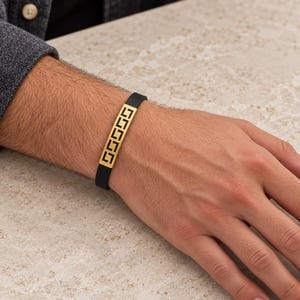 Könnte beinhalten: Ein schwarzes Silikonarmband mit einer mittigen, rechteckigen, goldfarbenen Metallplatte, die ein ausgeschnittenes griechisches Mäandermuster aufweist. Das Band ist glatt und flexibel und bildet einen Kontrast zum geometrischen Metalldesign.